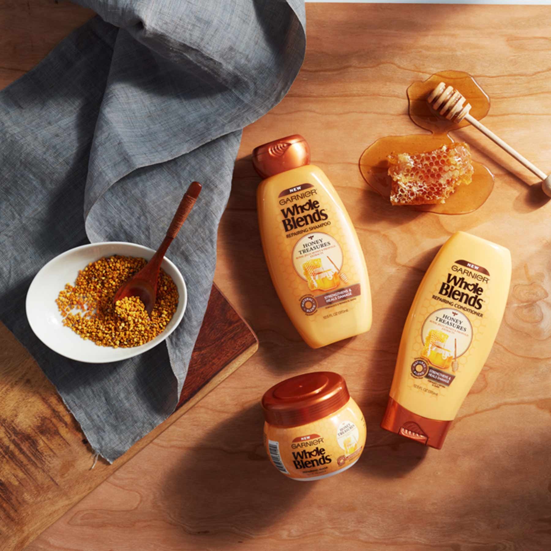 Whole Blends Honey Treasures Shampoo, Honey Treasures Conditioner, and Honey Treasures Repairing Mask on a wooden background next to a piece of dripping honey comb and honey dipper in a pool of honey, a wooden cutting board with a white bowl of bee pollen with a wooden spoon, and a blue-steel colored linen napkin.
